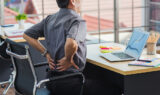 An Asian Businessman Sitting at a Desk Grabbing His Lower Back Dealing with Back Pain From Sitting at a Desk