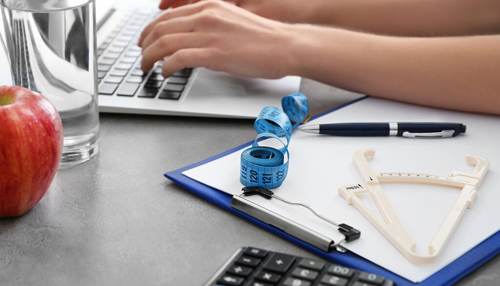 Closeup of a Nutritionist Typing on a Laptop With a Skin Catheter Measuring Tape & Clipboard Beside Them Who Qualifies for Gastric Sleeve Surgery