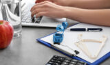 Closeup of a Nutritionist Typing on a Laptop With a Skin Catheter Measuring Tape & Clipboard Beside Them Who Qualifies for Gastric Sleeve Surgery