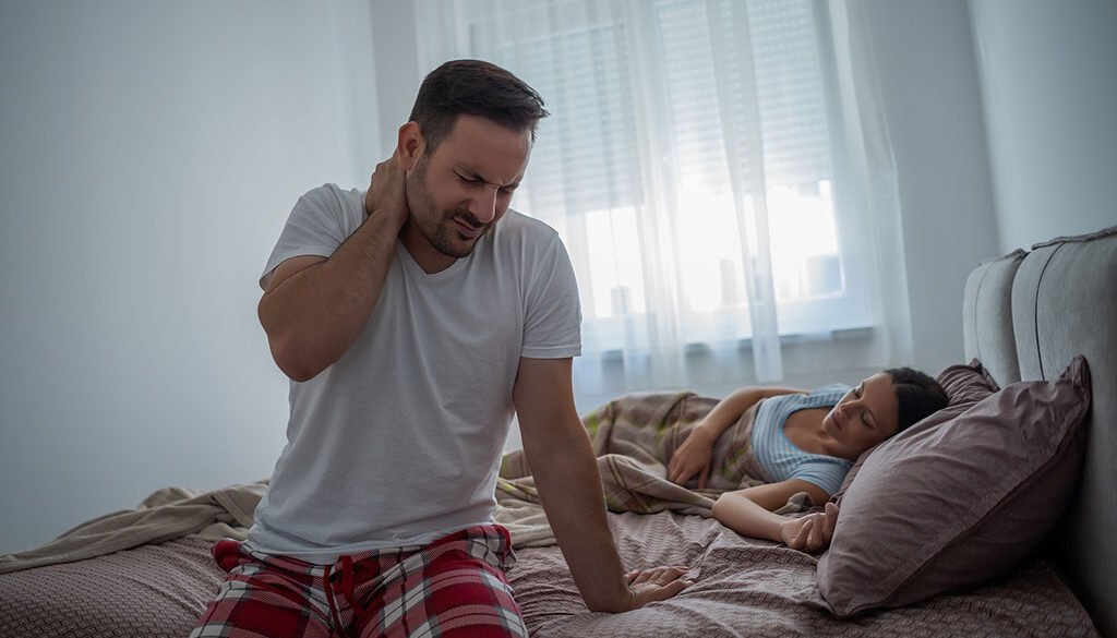 A Man Sitting on the Edge of a Bed Holding His Neck in Pain With His Partner Asleep Behind Him Neck Pain From Sleeping