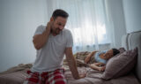 A Man Sitting on the Edge of a Bed Holding His Neck in Pain With His Partner Asleep Behind Him Neck Pain From Sleeping